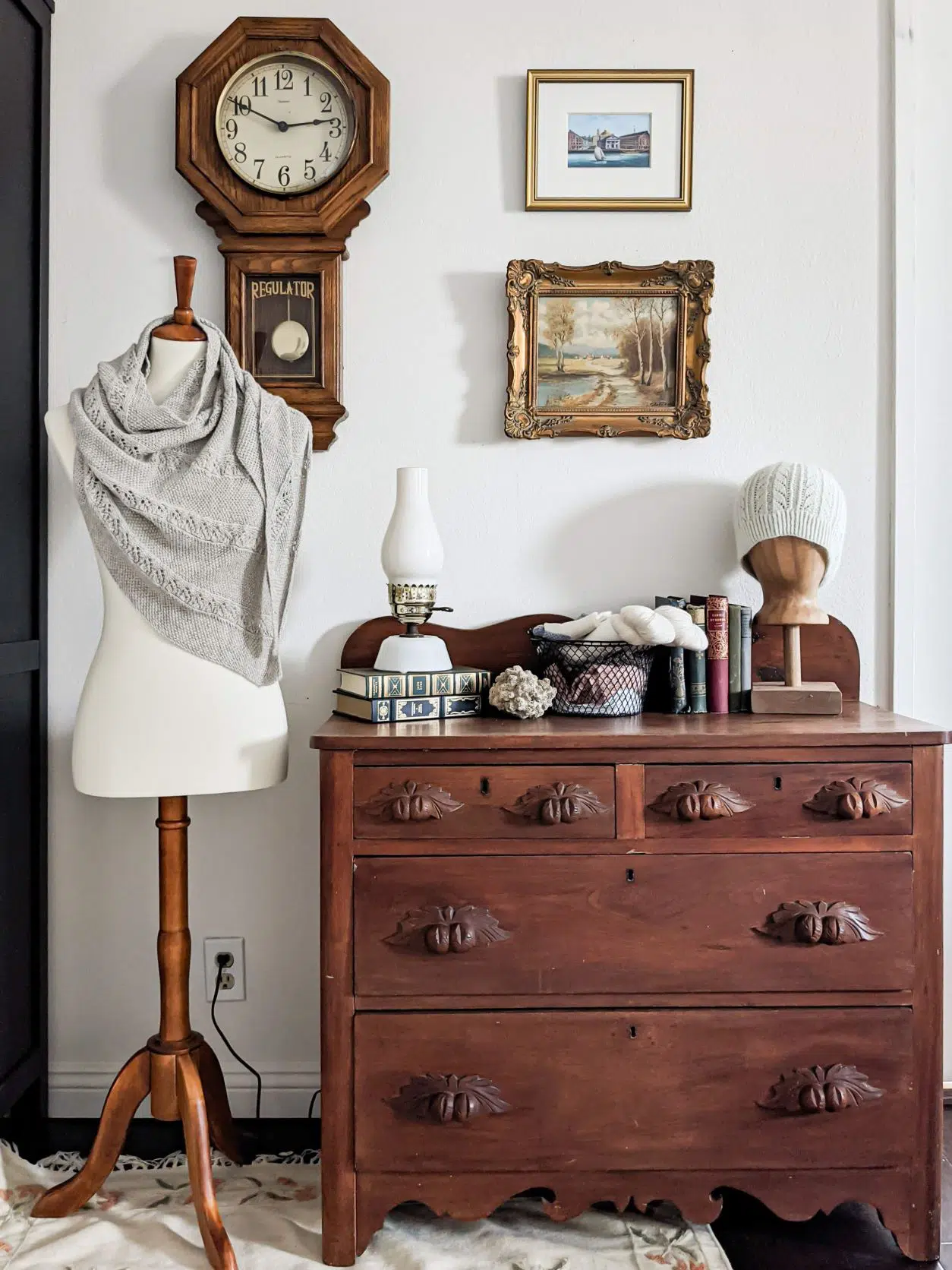 A small Eastlake dresser with carved handles sits next to a dressmaker's form with a gray shawl draped on it. Hung on the wall behind them are a wooden clock and two framed paintings. On top of the dresser are antique books, a lamp made to look like an oil lantern, a basket of yarn, and a wooden head form with a knit hat on it.