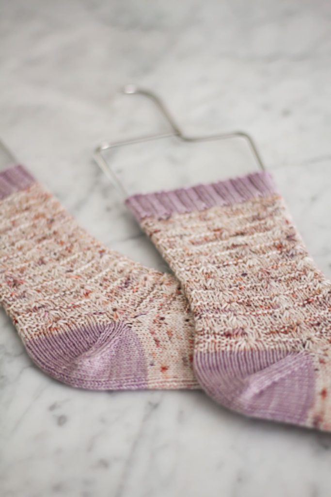 A pair of pink speckled socks with contrasting purple cuffs and heels are laid flat on a white surface. The image is zoomed in on the socks' peasant heel construction.
