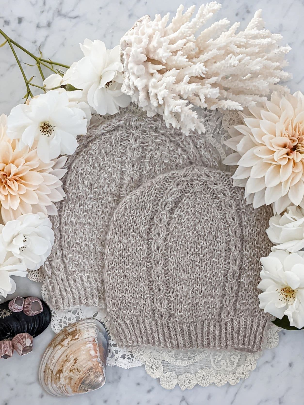 Two matching hats knit in fluffy tan yarn are laid flat on top of each other in a staggered fashion. They have seed stitch and columns of coin lace. They're surrounded by peach dahlias, white roses, two seashells, and spiky white coral.