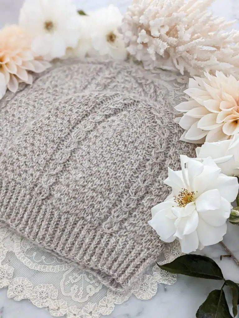 Two matching hats knit in fluffy tan yarn are laid flat on top of each other in a staggered fashion. They have seed stitch and columns of coin lace. They're surrounded by peach dahlias, white roses, and spiky white coral.
