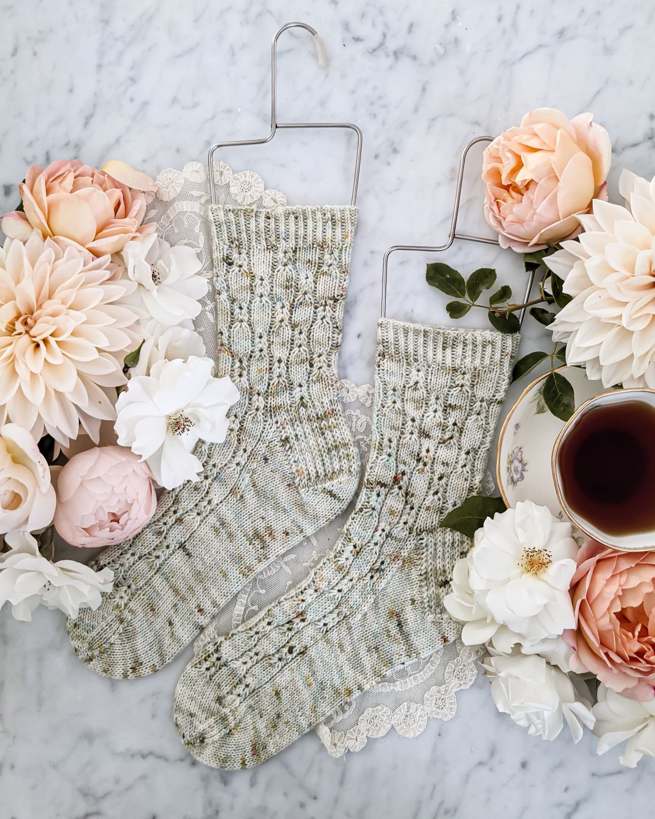 A pair of speckled, pale green socks with columns of eyelet details is laid flat on a white marble surface. Both toes point to the left. They are surrounded by creamy dahlias, peach and white roses, and a white teacup filled with tea.
