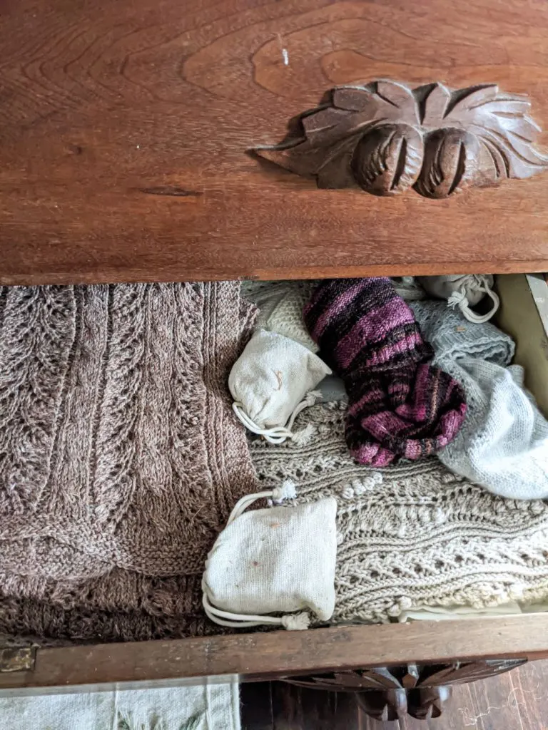 A peek inside a dresser drawer where knitwear is stored. There are tan and brown scarves with lacy textures, two pairs of socks folded together (one purple striped, one gray), and two sachets full of cedar chips. Visible at top right is a carved wooden handle on the drawer above the one that's open.