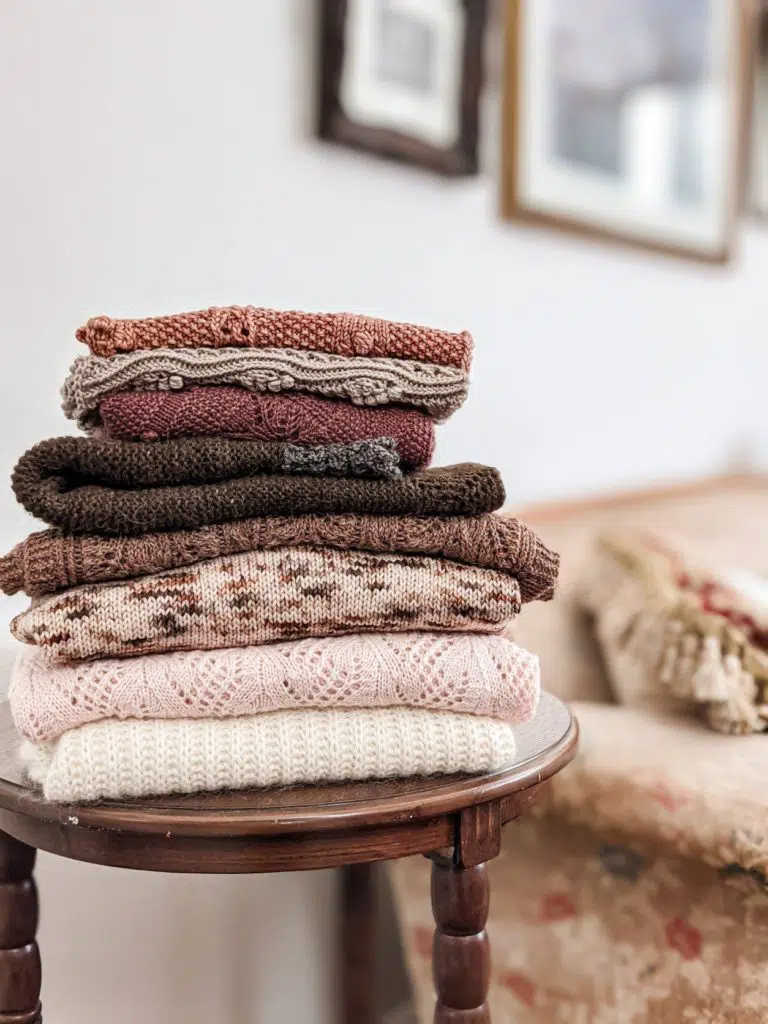 A folded pile of cream, pink, speckled, tan, and chocolate brown knitwear on top of a brown wooden table.