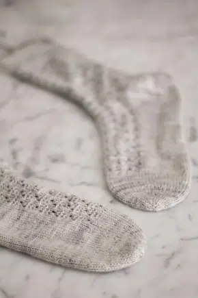 A pair of pale blue-gray socks with round toes are laid flat on a white marble surface. The toes point toward each other.