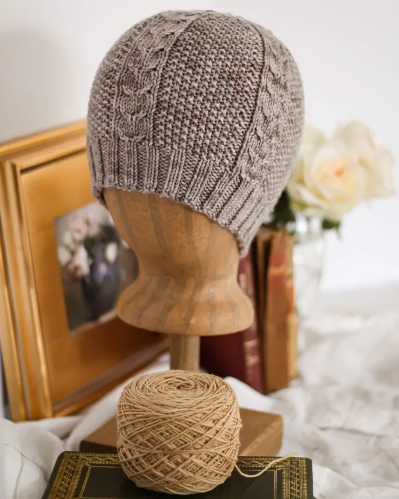 A gray knit hat with cabled columns and seed is on a wooden head form to show what the hat would look like on a person. In the foreground is a green book with a gilded cover and a cake of tan yarn. In the background, blurred out, are some framed artwork, antique books, and white roses in a glass vase..