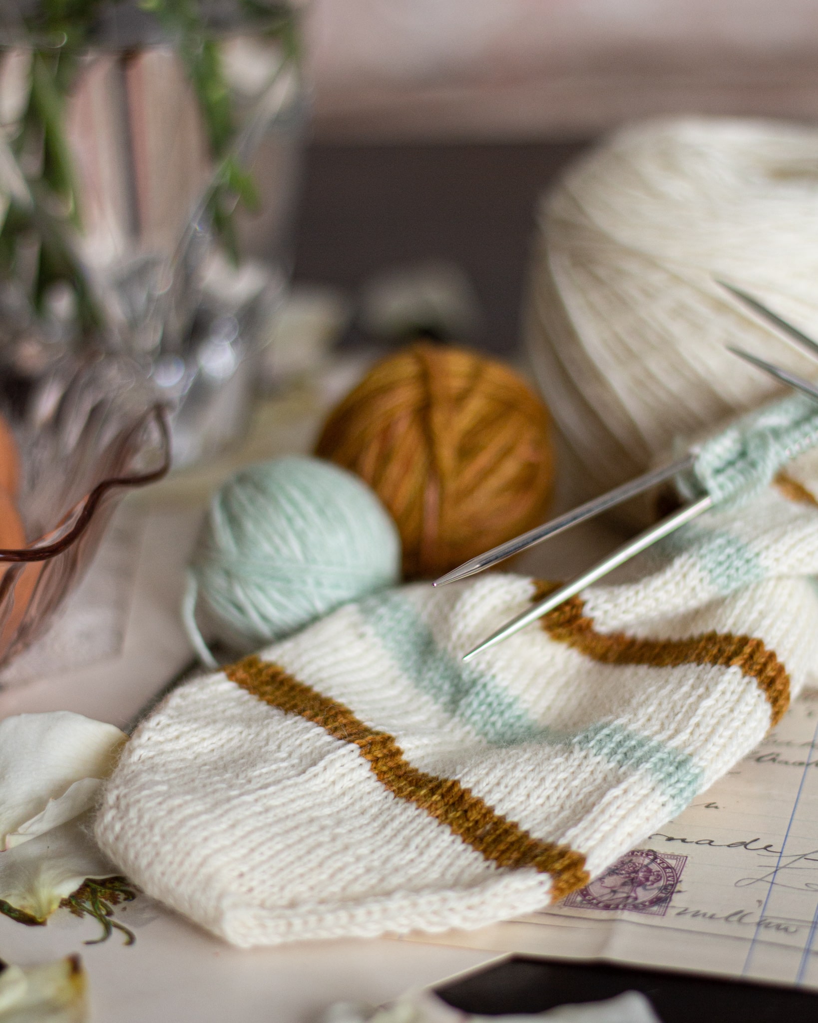 A white, caramel, and mint striped sock toe is laid flat with small balls of mint and caramel yarn behind it. The sock is knit on double-pointed needles.