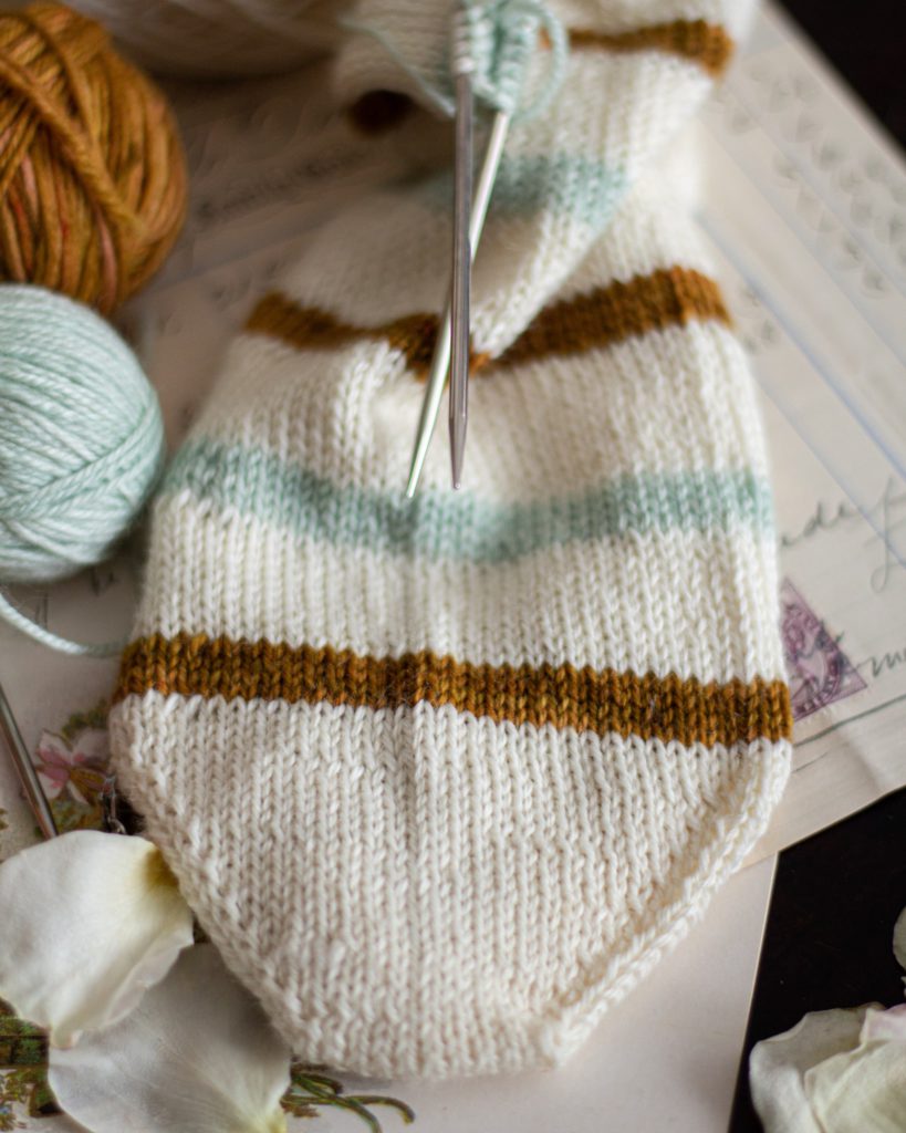 A white, caramel, and mint striped sock toe is laid flat with small balls of mint and caramel yarn next to it. The image is zoomed in on the tip of the toe for a better look at how the increases work.
