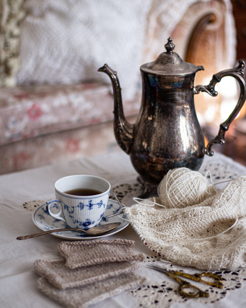 In the foreground are ornate brass scissors and three tan swatches of knit fabric. The middle distance in focus features a pile of creamy white knitting, a blue and white teacup and saucer, and a silver coffee pot. Blurred in the background is a pale floral sofa.