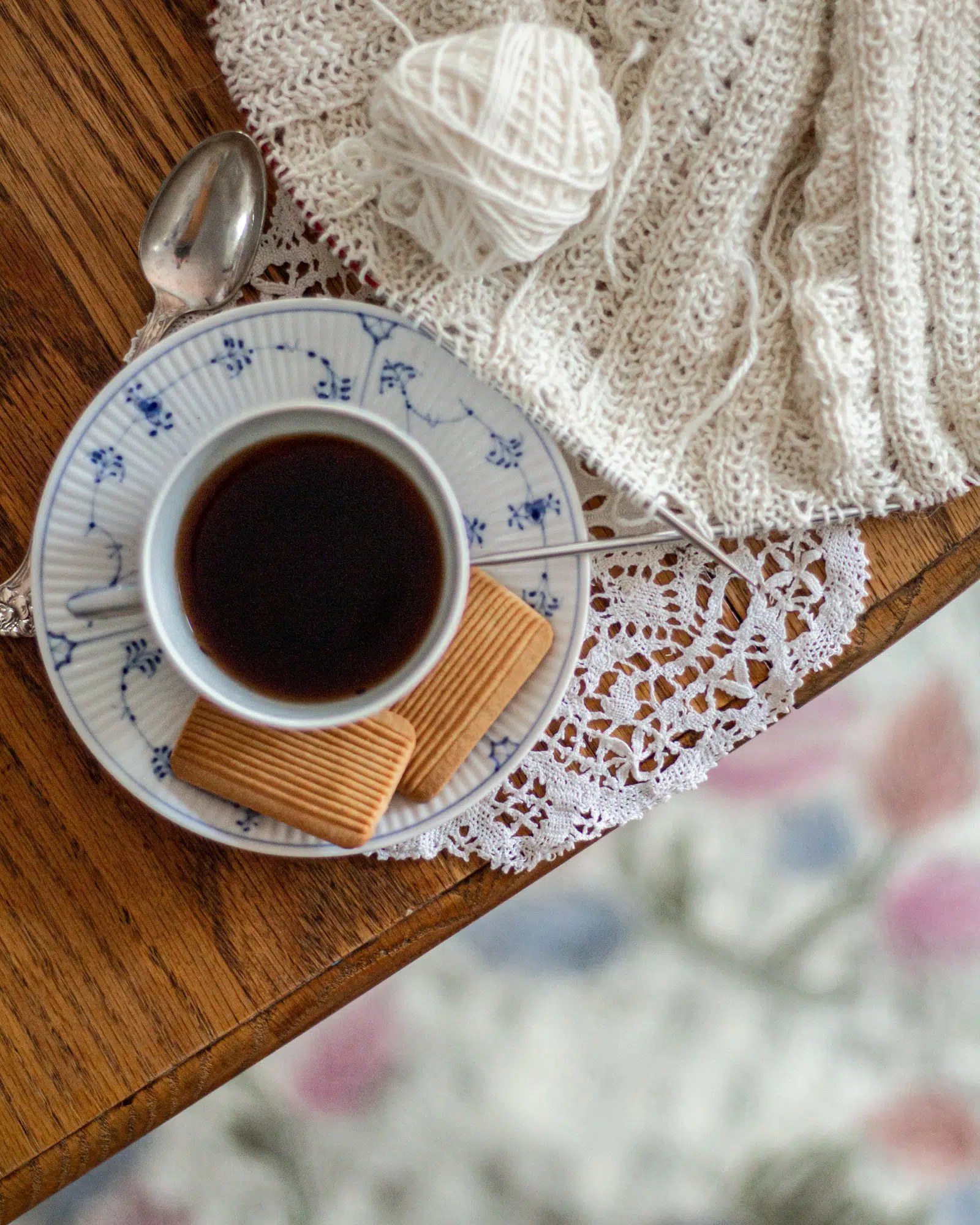 A top-down shot of a blue and white teacup and saucer sitting on the edge of an oak table with a white doily and some white knitting on steel needles. Knitting helps me overcome perfectionism by giving me a safe place to make mistakes.