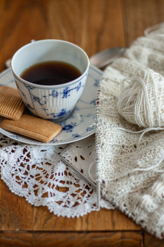 A sideways shot of a blue and white teacup and saucer (on the left) sitting on an oak table with a white doily and some white knitting on steel needles. Knitting helps me overcome perfectionism by giving me a safe place to make mistakes.