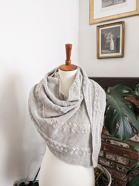 An asymmetrical triangle-shaped shawl in a soft gray color is draped around the torso of a white dressmaker's form. In the background is a large monstera plant, a brick fireplace, and some framed antique prints on the walls.