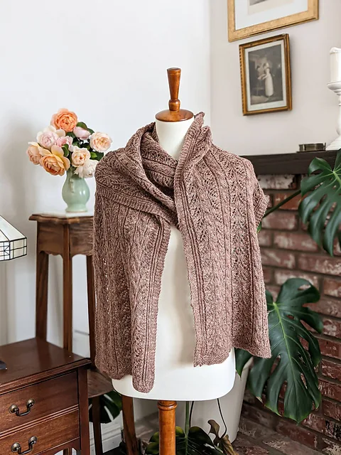 A rectangle-shaped shawl in a chocolate brown color is draped around the torso of a white dressmaker's form. In the background is a large monstera plant, a brick fireplace, a vase filled with various sherbet-colored roses, and some framed antique prints on the walls.