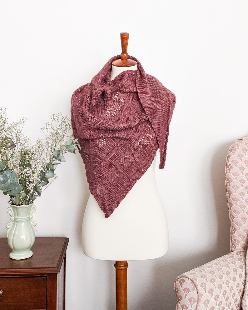 The Berry Scone Shawl, an asymmetrical triangle with stripes of lace, bobbles, and seed stitch, is draped around a white dressmaker's form. It's knit up in a desaturated burgundy color that's reminiscent of baked berries.