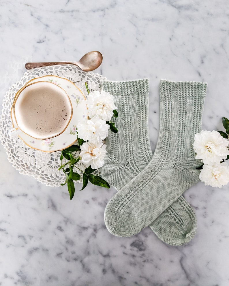 The Tiramisu Socks, a pair of mint green socks are laid flat on a white marble countertop with the toes pointing inward. They're surrounded by white roses and a white teacup full of a frothy latte.