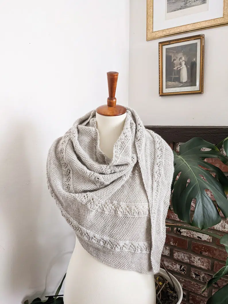 The Plenitude Shawl, a gray, asymmetrical triangle with columns of lace and seed stitch, is draped around the shoulders of a white dressmaker's form.
