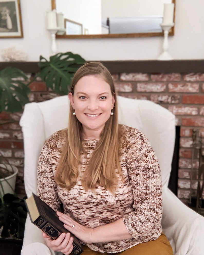 A picture of me sitting in a white armchair wearing my Geniality Sweater, which is knit in a cream-colored yarn with brown and burgundy speckles. I have long blonde hair, am holding a book in my right hand, and am smiling at the camera.