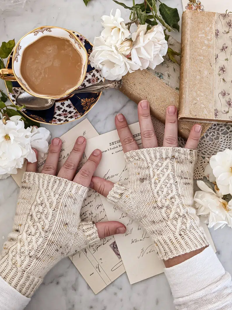 A pair of cream-colored fingerless mitts with cables up the backs are worn by two small, plump hands, surrounded by white roses, antique books, and a navy blue teacup full of coffee.