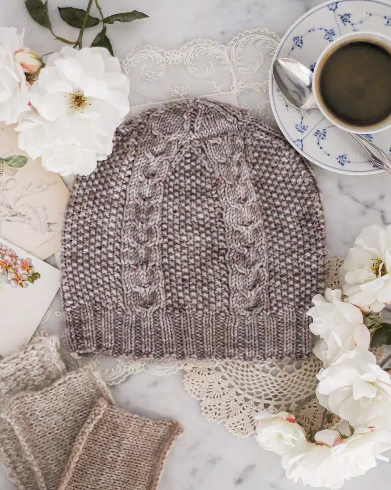 A gray knit hat with cabled columns and seed stitch sits flat on a white marble countertop, surrounded by a teacup full of coffee, white roses, swatches of tan knit fabric, and antique paper ephemera.