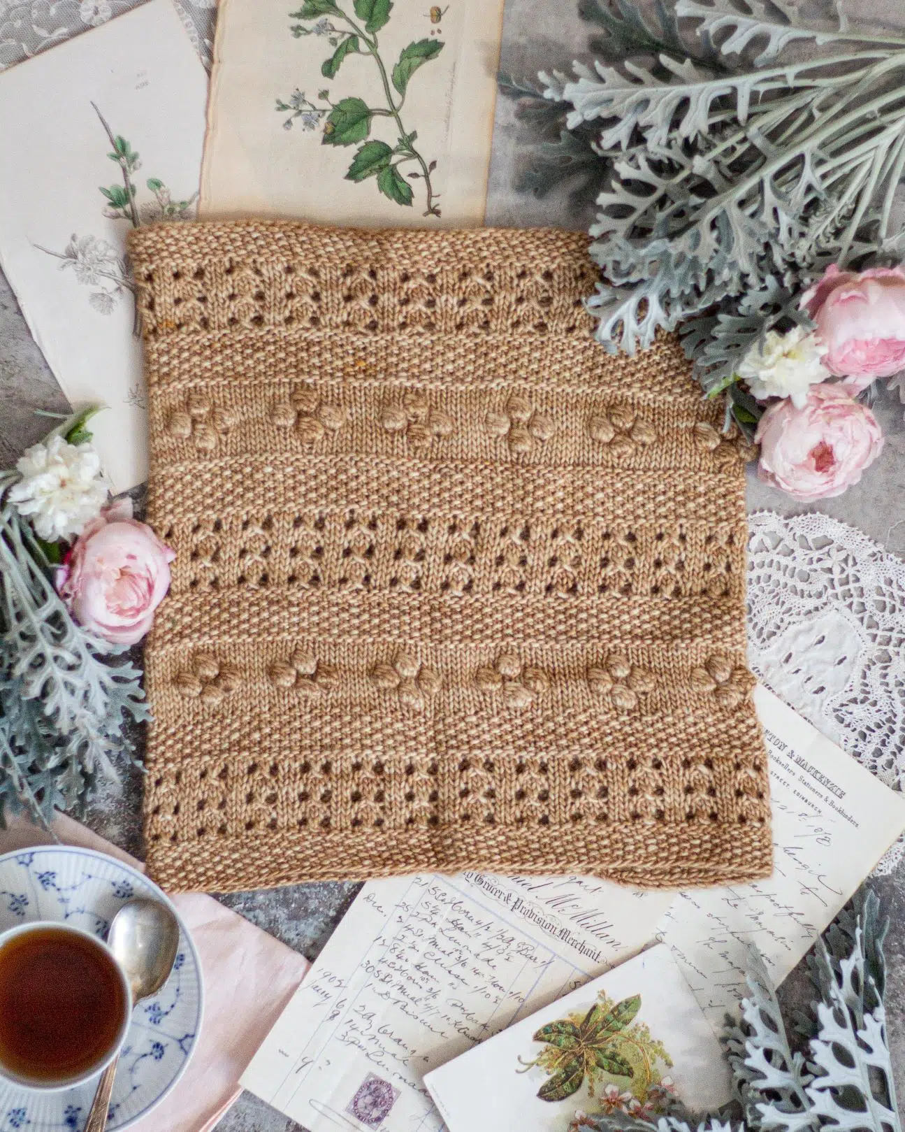 A golden-colored cowl with alternating rows of eyelets, bobbles, and seed stitch is laid flat and surrounded by pink and white roses, antique paper ephemera, and a blue and white teacup. The photo is shot from the top straight down.