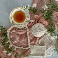 A top-down photo of cream-colored yarn in a ball with some swatches and a new project just cast on to circular needles. The yarn is all laid out on top of a deep pink gauze fabric on a gray background. To the top left are a teacup and saucer, and there are several sprigs of greenery at the edges.