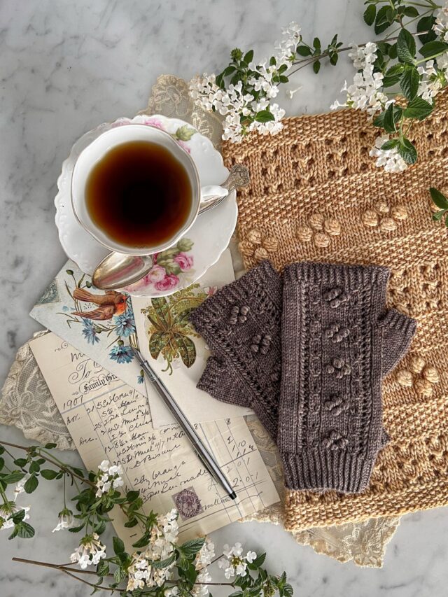 A top-down shot of a pair of brown fingerless mitts resting on top of a golden cowl, both adorned with many bobbles. To the side are antique paper ephemera and a pink and white teacup.