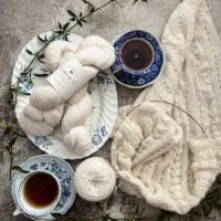 A top-down view of a lacy white wrap, half-knitted and still on the needles. To its left are two blue and white teacups filled with tea, along with two more skeins of white yarn on a blue and white platter.