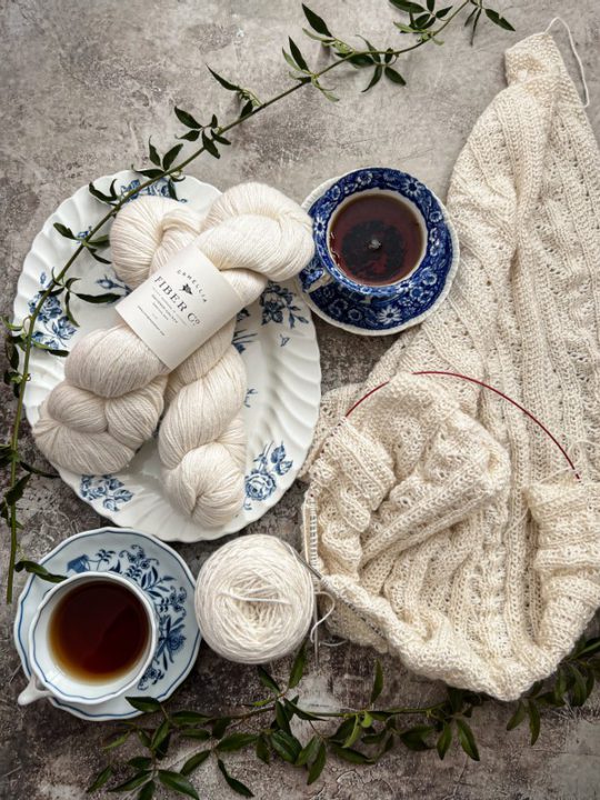 A top-down view of a lacy white wrap, half-knitted and still on the needles. To its left are two blue and white teacups filled with tea, along with two more skeins of white yarn on a blue and white platter.