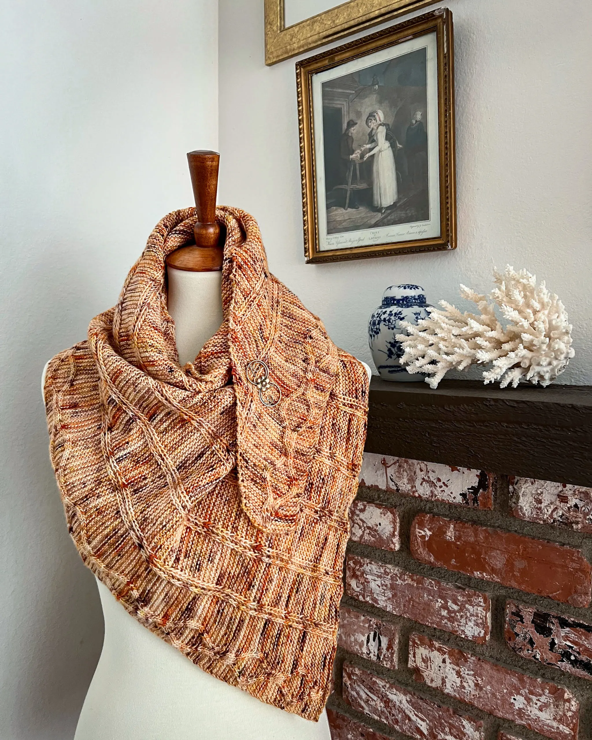 An orange triangular shawl with columns of slipped stitches and a cabled edging is wrapped around a white dressmaker's form. In the background are a brick fireplace, some white coral, a blue and white ginger jar, and antique prints on the wall.