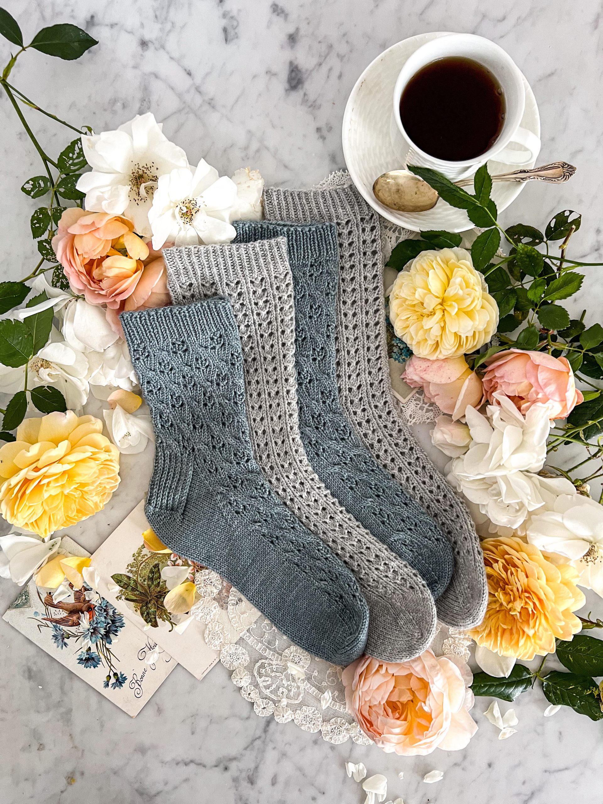 Four socks, stacked on each other in alternating blue and gray, photographed straight on. They're surrounded by yellow, white, and pink roses, a teacup full of coffee, doilies, and antique postcards..