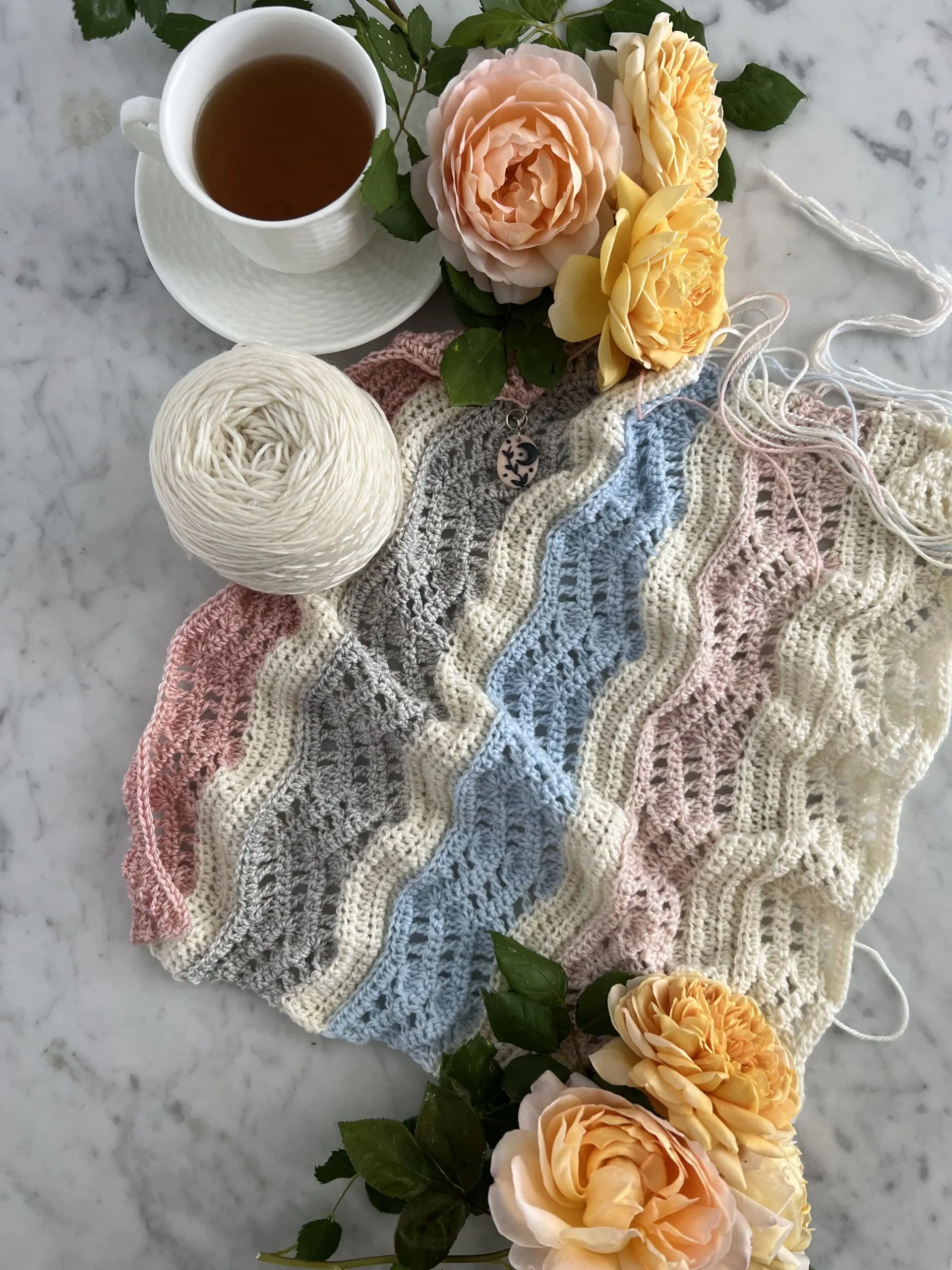 A ripply crocheted wrap in progress. It has stripes in shades of cream, pink, blue, and gray. There are also orangey-yellow roses and a white teacup full of tea.