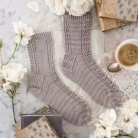 A top-down image of the Lucida Socks, a pair of lavender-colored socks with panels of eyelets and cables running down front and back, modeled on a pair of sock blockers and laid flat on a white marble background surrounded by antique books, which roses, and a teacup full of espresso