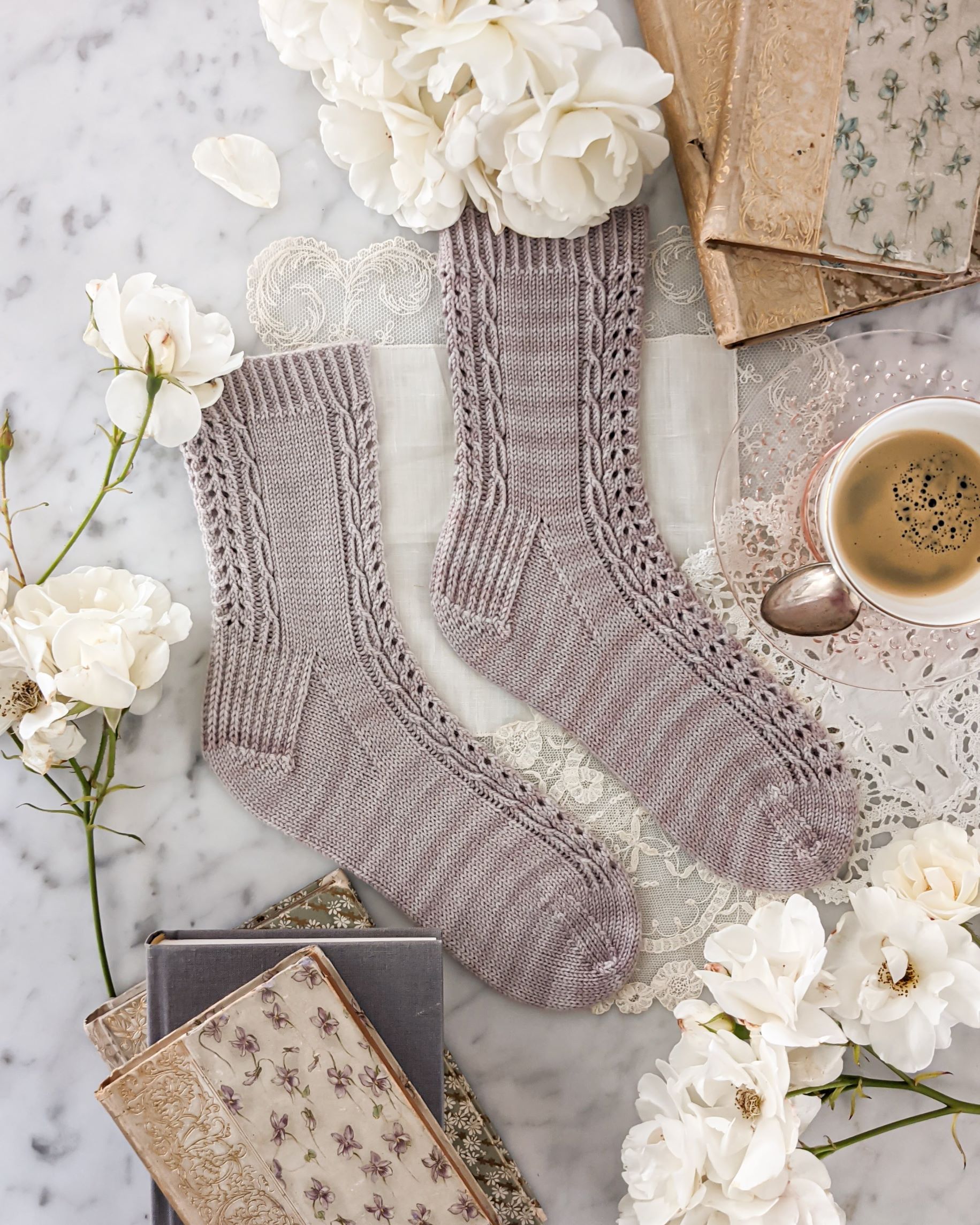A top-down image of the Lucida Socks, a pair of lavender-colored socks with panels of eyelets and cables running down front and back, modeled on a pair of sock blockers and laid flat on a white marble background surrounded by antique books, which roses, and a teacup full of espresso
