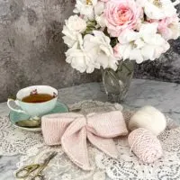 A pink, fuzzy, knit bow sits in the center of the photograph, surrounded by a vase of pink and white roses, some small balls of yarn, and a polka dotted teacup