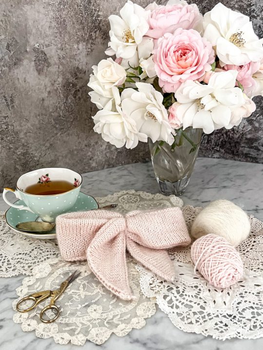 A pink, fuzzy, knit bow sits in the center of the photograph, surrounded by a vase of pink and white roses, some small balls of yarn, and a polka dotted teacup