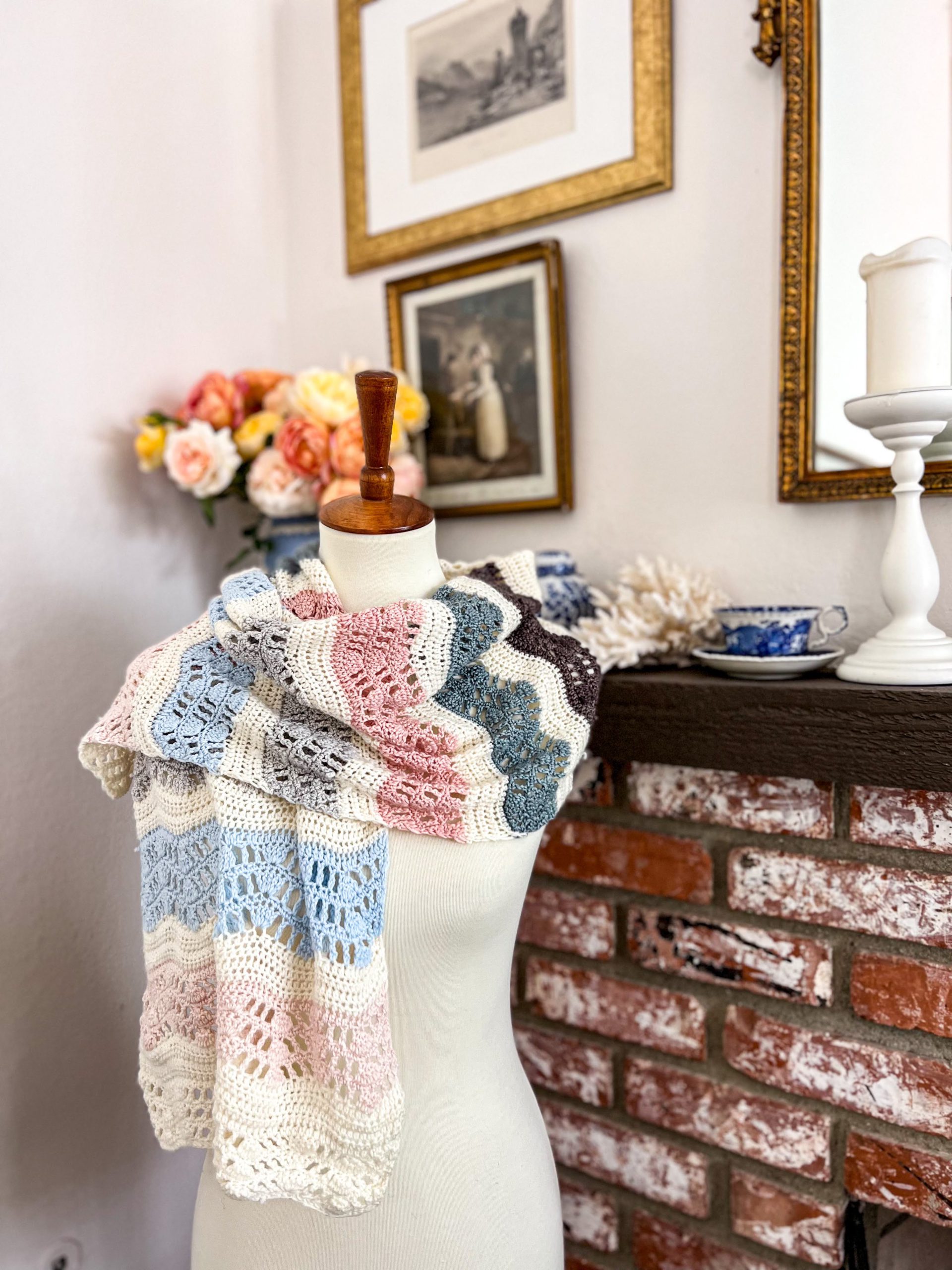 A rippled, lacy, crocheted wrap with stripes in various shades of white, blue, and pink is draped around the shoulders of a white dressmaker's form.