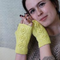 A young, white woman with dark brown hair rests her cheek against her hands, which are wearing a pair of yellow fingerless mitts with cables and bobbles running up the back.