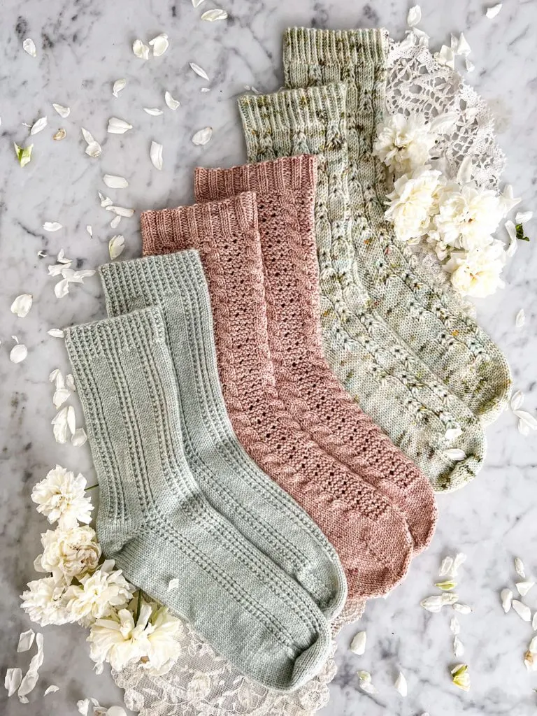 Three pairs of socks—one mint green, one pink, and one green with brown and yellow speckles—are laid out in a staggered stairstep fashion on top of a white marble countertop. They're surrounded by small white roses.