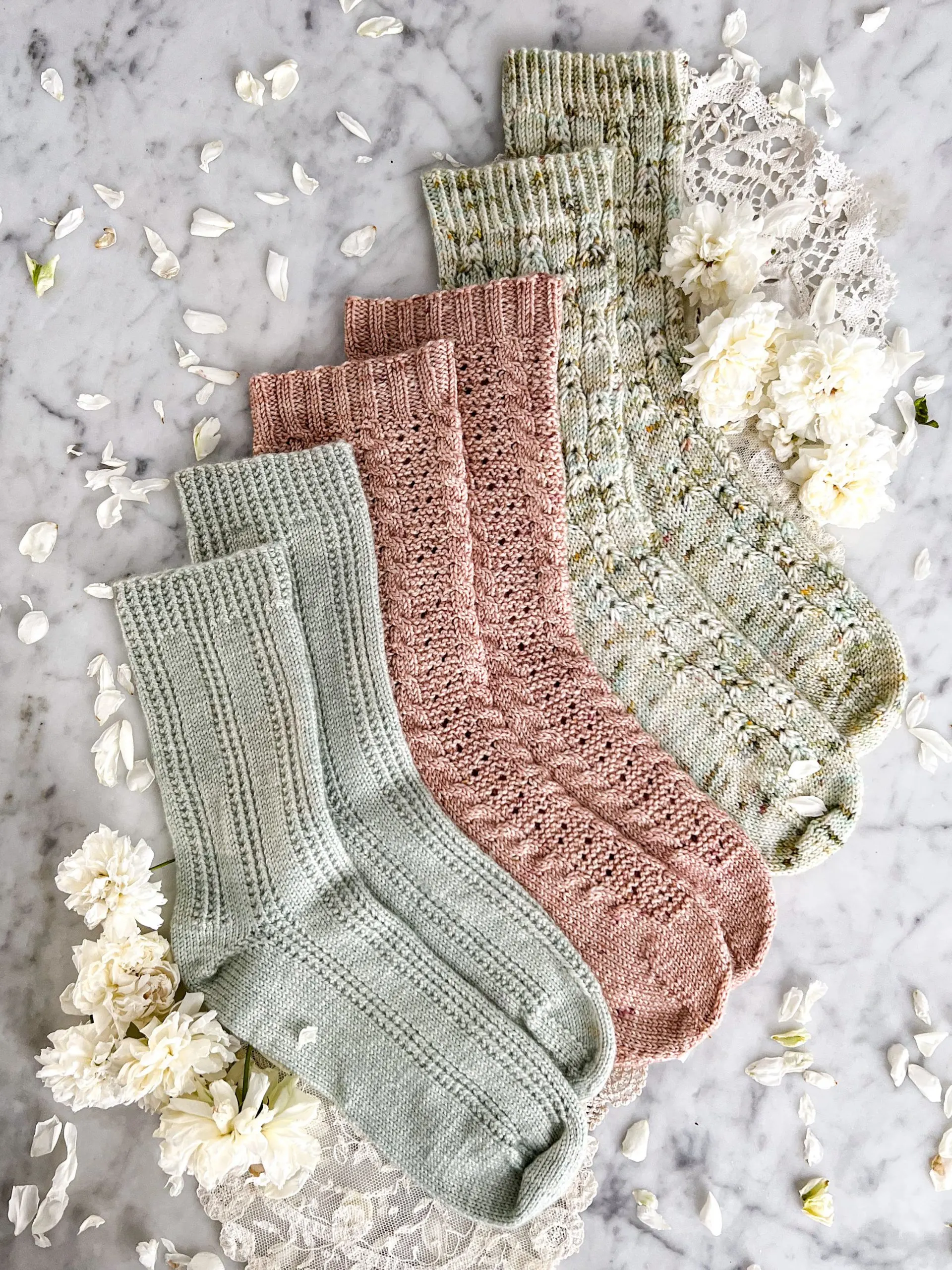 Three pairs of socks—one mint green, one pink, and one green with brown and yellow speckles—are laid out in a staggered stairstep fashion on top of a white marble countertop. They're surrounded by small white roses.