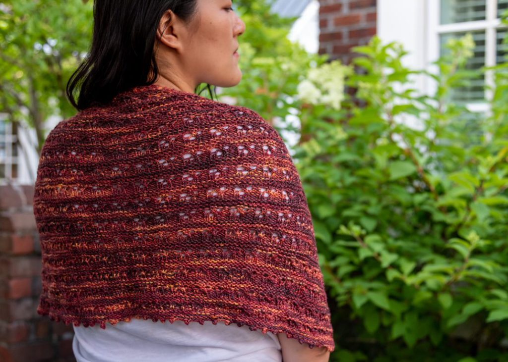 The Dusk in the Desert Shawl, a red shawl knit in a semicircular shape, is draped around the shoulders of a woman with long, black hair. She is photographed from the back and looking to her right.