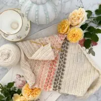 A crocheted shawl in shades of cream, green, and several pink varieties is laid out, slightly rumpled, on a white marble surface. It's surrounded by a teapot with a texture that looks like a sea urchin, an empty teacup, several yellow roses, and a couple antique rose botanical prints.