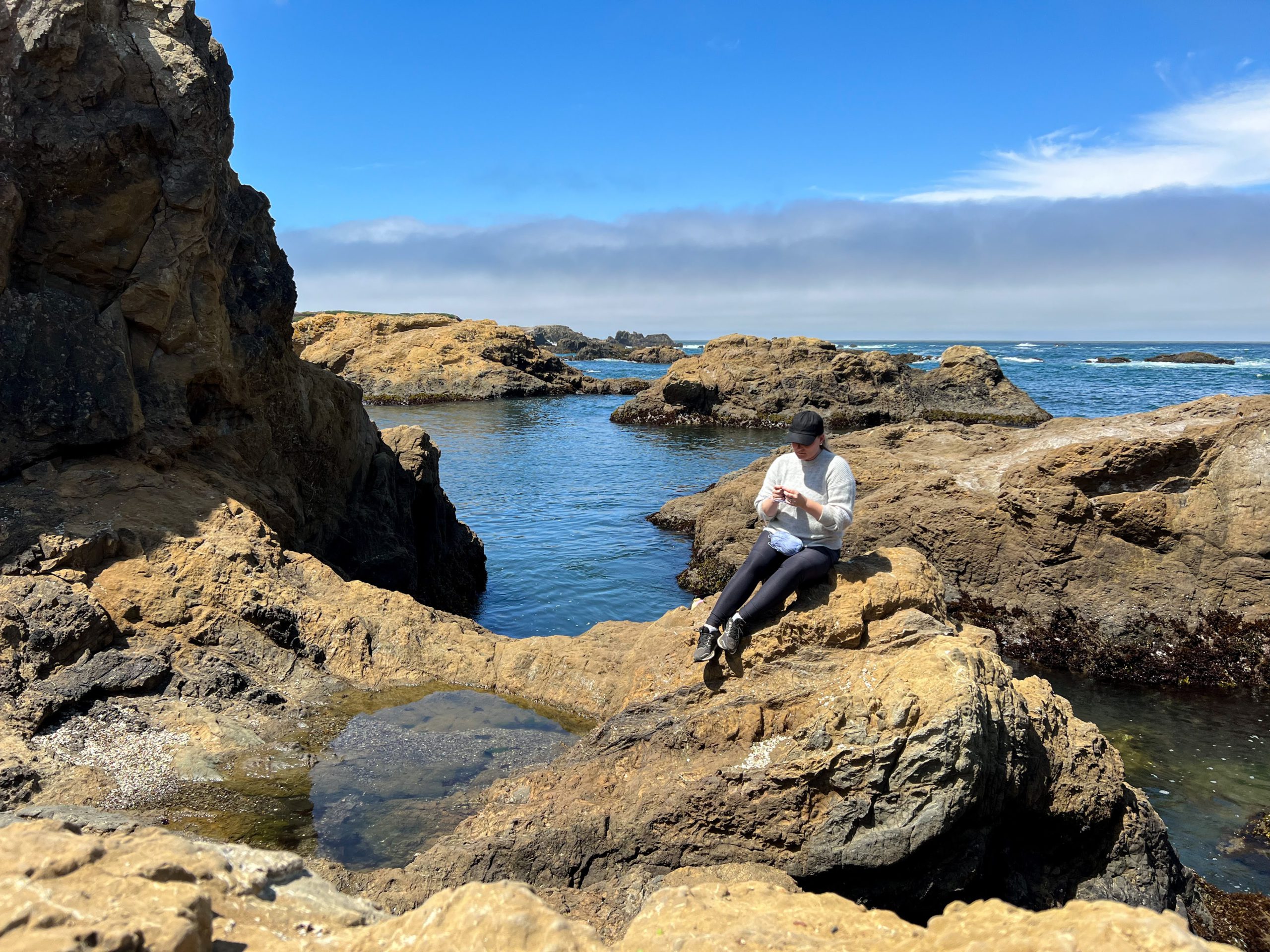 A woman in a black baseball cap, gray sweater, and black leggings sits knitting on a rocky outcropping with the ocean and some dramatic, looming clouds behind her.