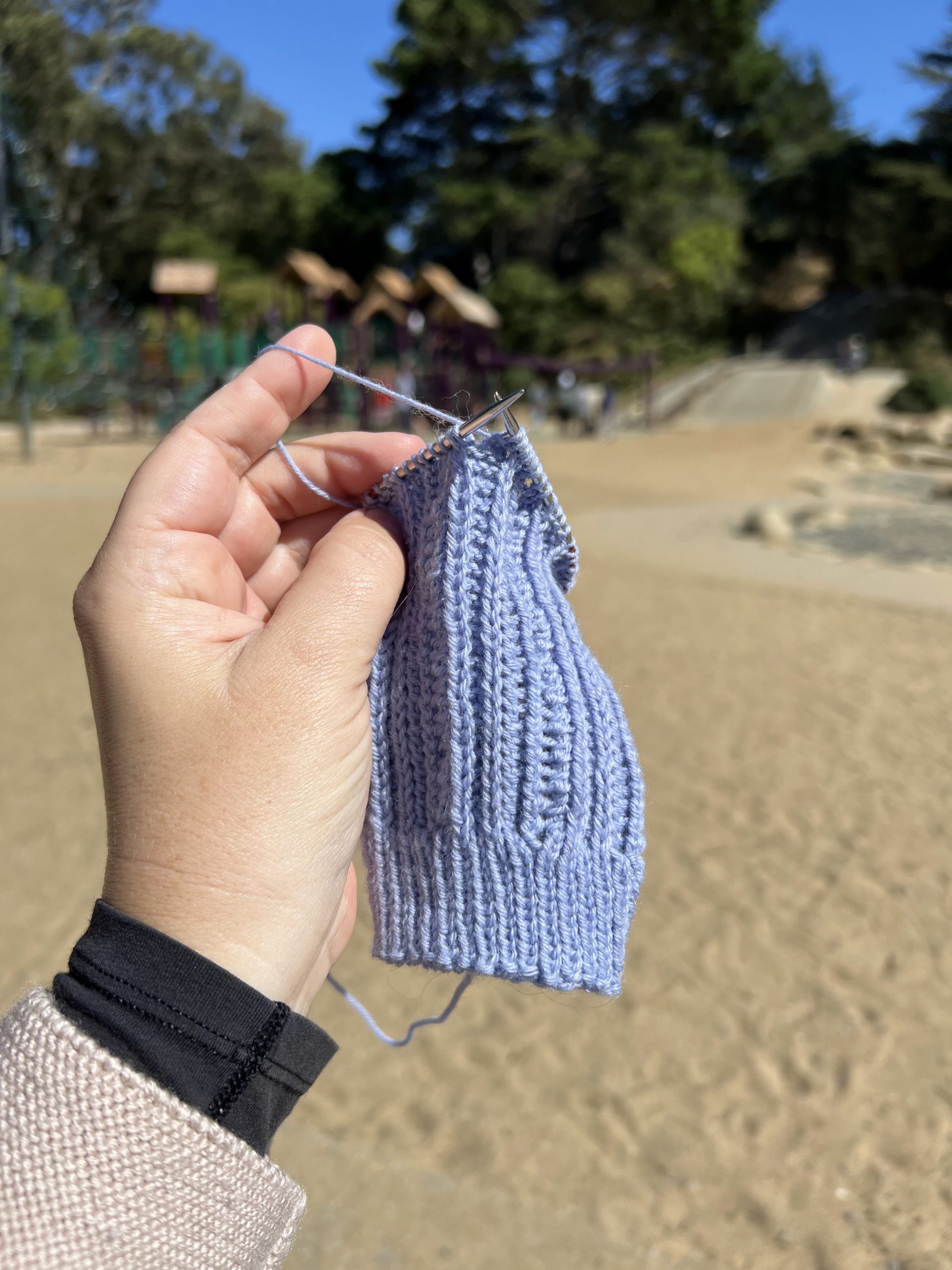 A small, plump hand holds up a half-knit sock leg. Blurred in the background is lots of sand and a playground structure.