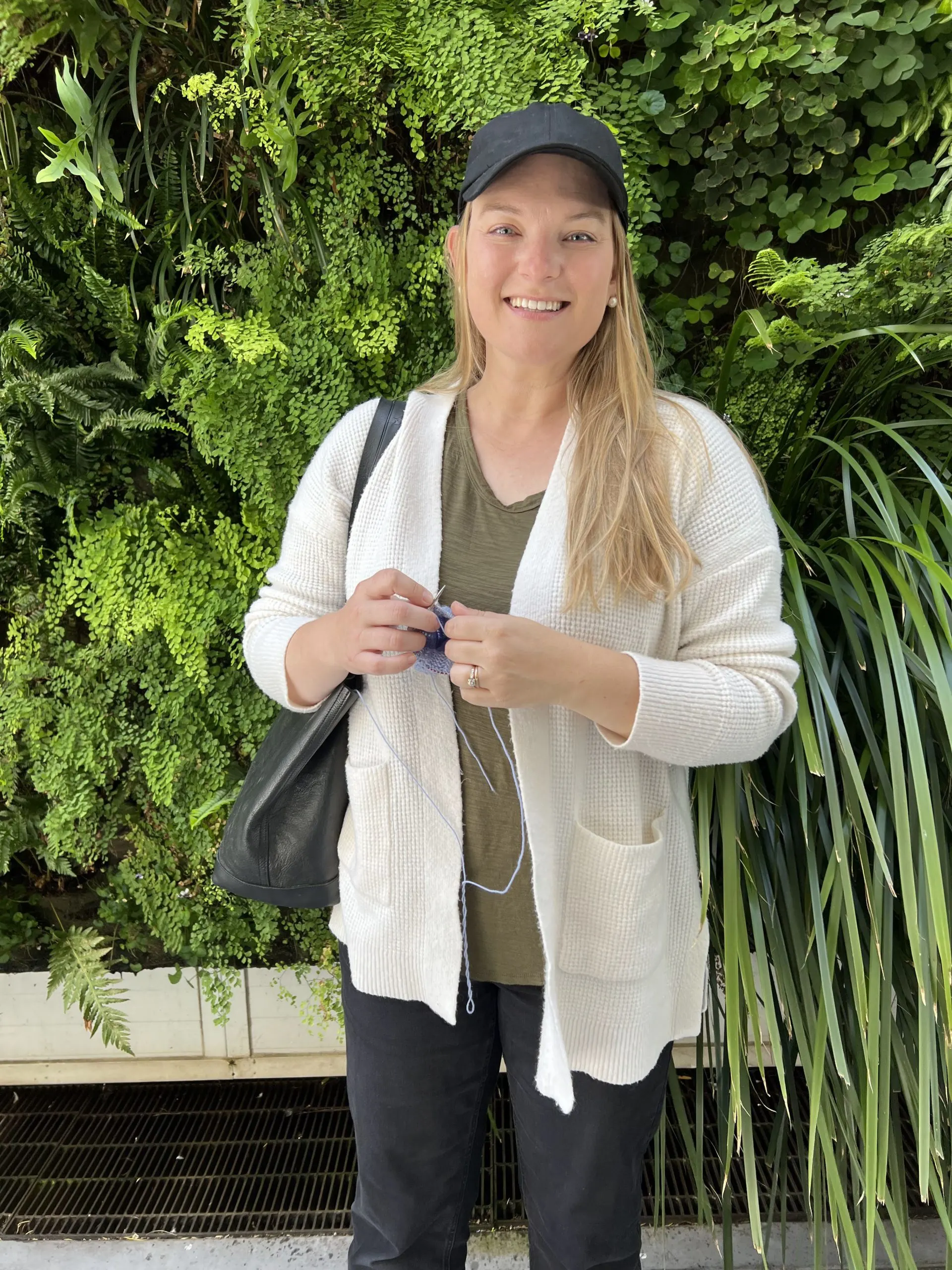 A late-30s woman with long blonde hair and a white cardigan stands, knitting, in front of a plant wall featuring 37 varieties of plant life.