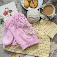 A top-down image of a pale yellow crocheted child's sweater with a pink knit child's sweater folded on top and a cream-colored ball of yarn. They're surrounded by a teacup full of coffee, a plate with tiny baked treats, and antique paper ephemera.