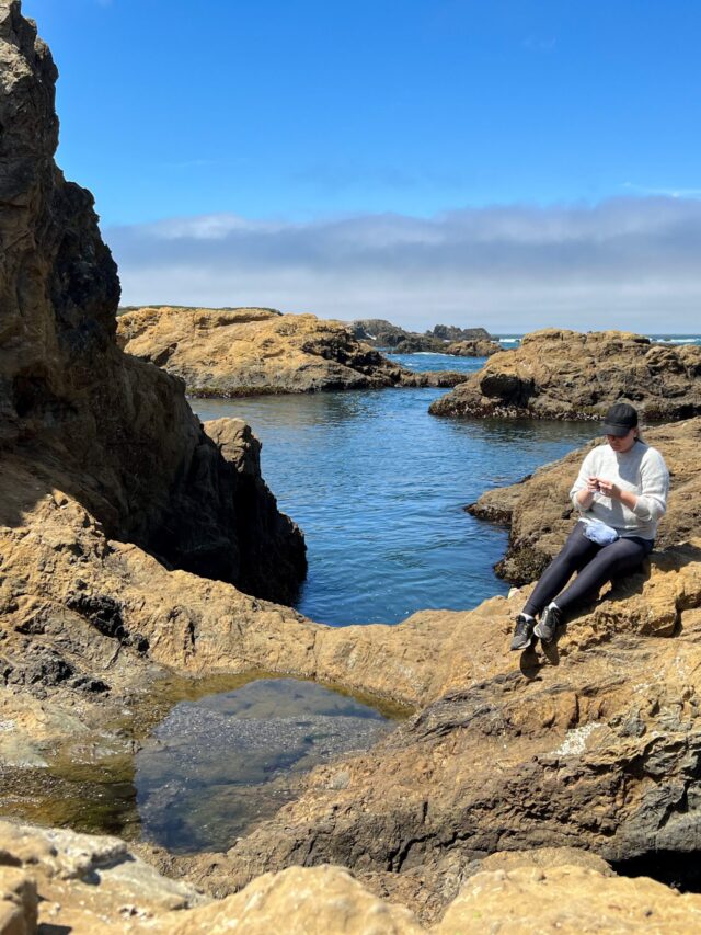 A woman in a black baseball cap, gray sweater, and black leggings sits knitting on a rocky outcropping with the ocean and some dramatic, looming clouds behind her.