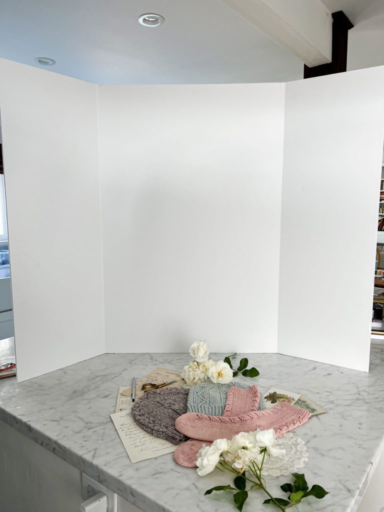 A large, white, folding foam board sits on top of a white marble countertop. In front of it is a flatlay of various pastel knit items and paper ephemera.