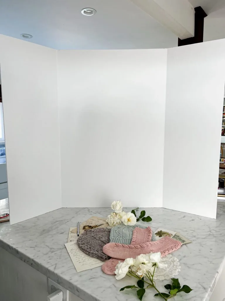 A large, white, folding foam board sits on top of a white marble countertop. In front of it is a flatlay of various pastel knit items and paper ephemera.