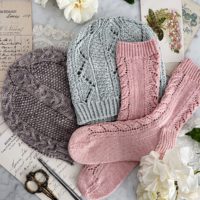 A top-down image of pastel knit items - a gray hat, a light blue hat, and pink socks - surrounded by paper ephemera and white roses on top of a white marble countertop.