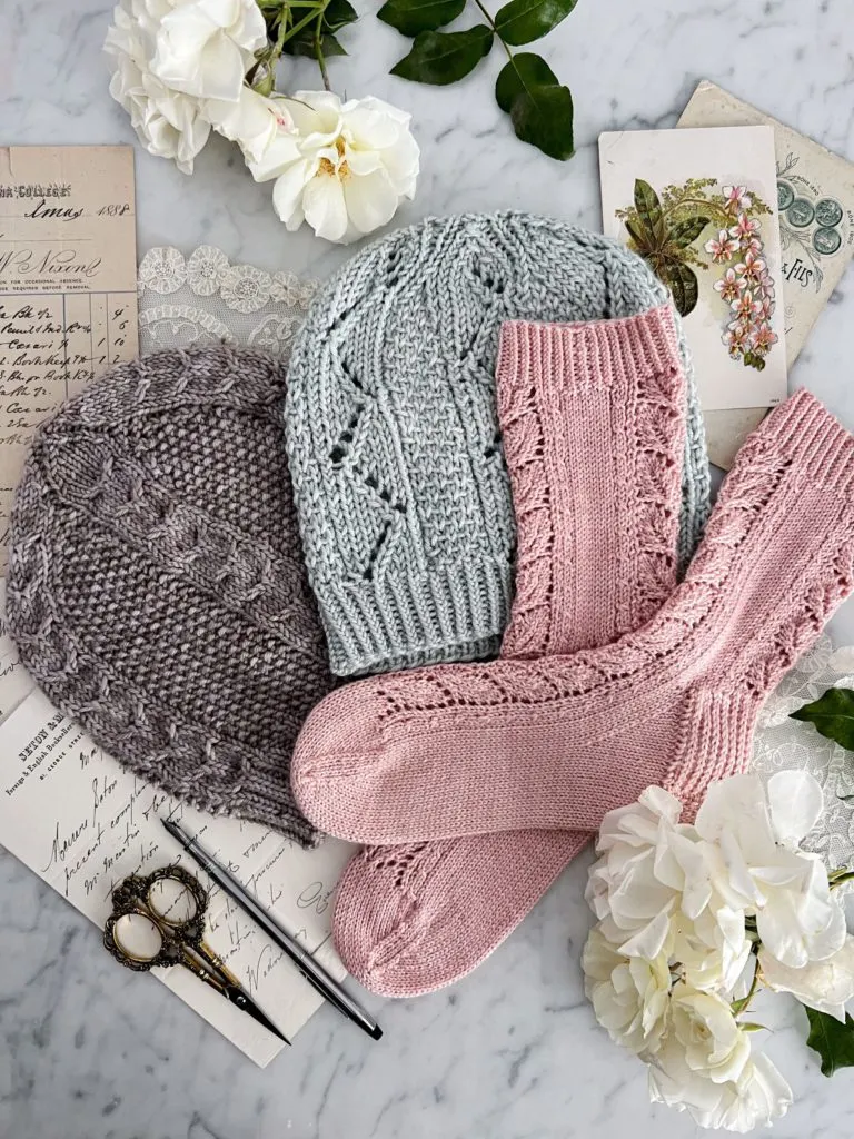 A top-down image of pastel knit items - a gray hat, a light blue hat, and pink socks - surrounded by paper ephemera and white roses on top of a white marble countertop.