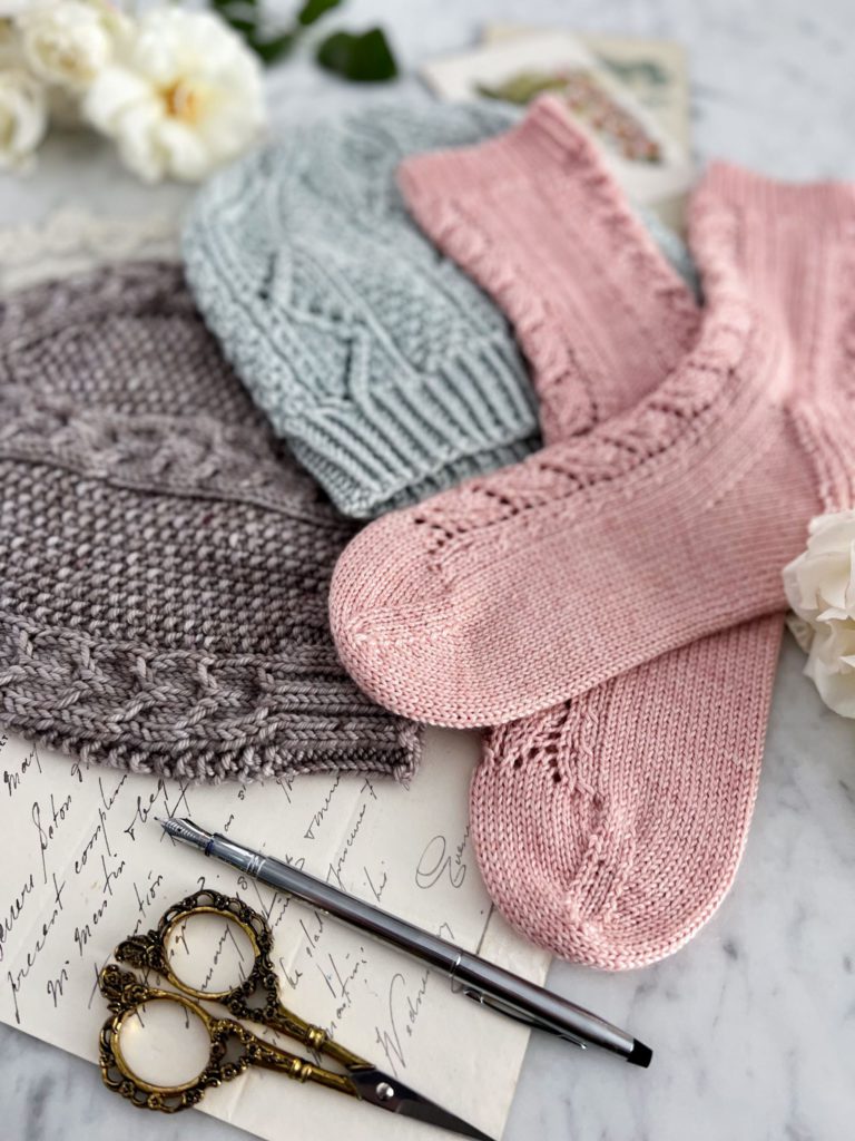 A close-up on the toes of some pink, handknit socks, which are laid out on top of a light blue handknit hat and a gray handknit hat. In the foreground are some ornate scissors and a silver fountain pen.